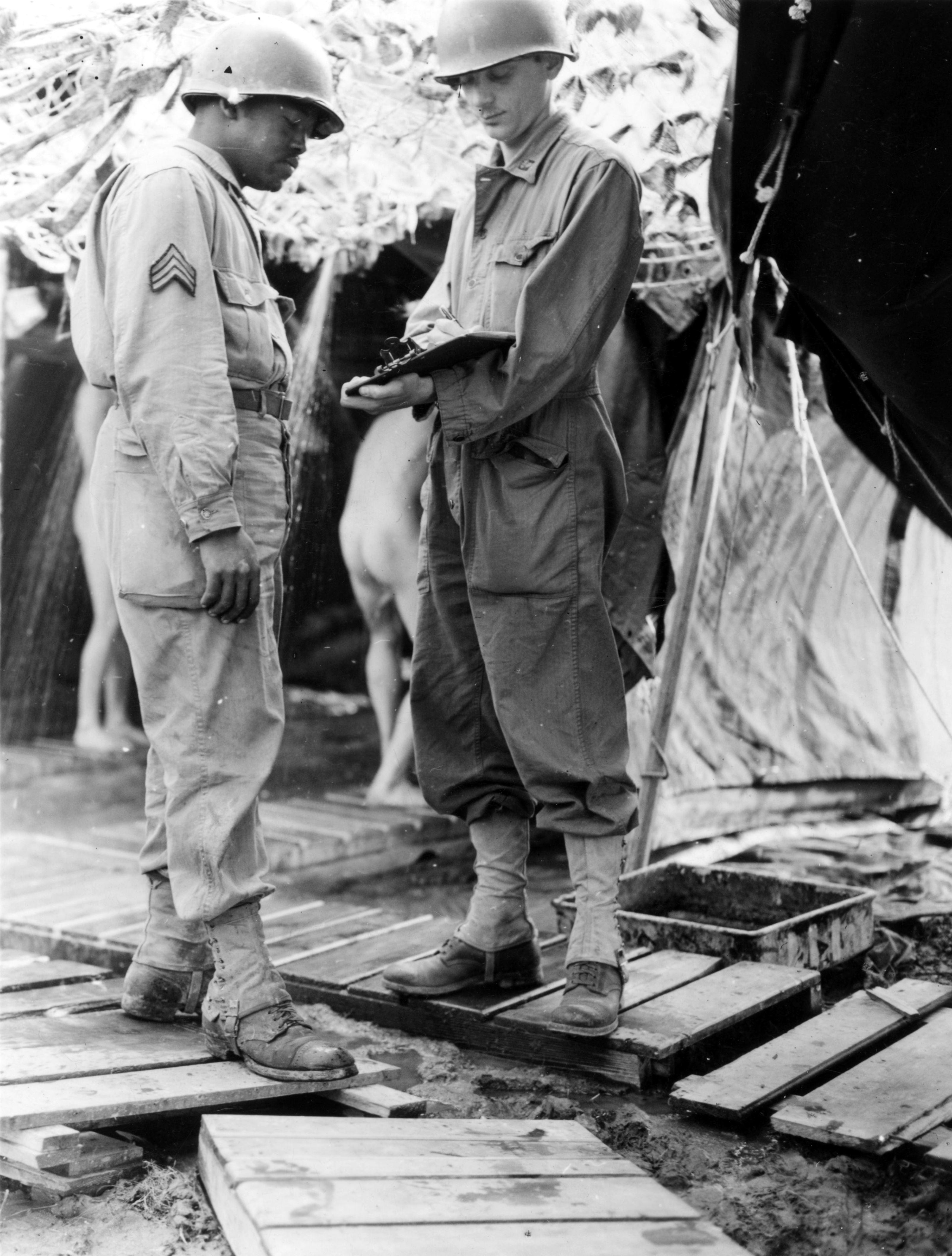 Historical image showing Sgt. John B. Smith and Lt. Robert Back check records in front of showers for front line troops. of Black American soldiers during WWII